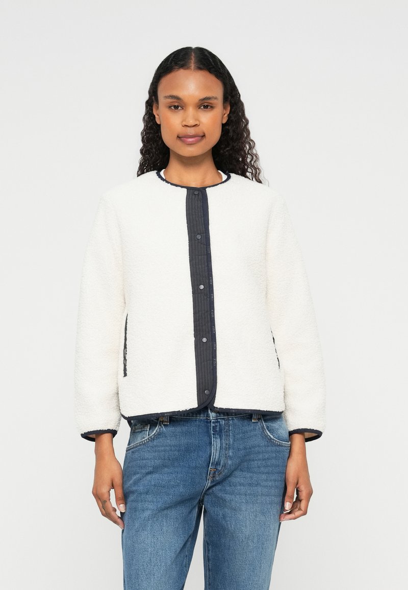 Veste en polaire blanche avec col rond, patte de boutonnage bleu marine et poches latérales. Caractéristiques d'un tissu texturé et d'un bord bleu marine contrastant.