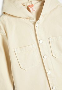 Beige denimjakke med hætte, udstyret med hvide trykknapper og to frontlommer. Glat tekstur med synlige syninger.
