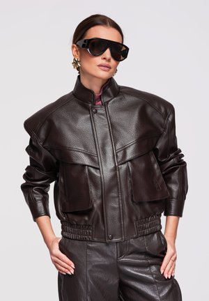 Femme portant une veste et un pantalon en cuir noir oversized, de grandes lunettes de soleil noires et des boucles d'oreilles géométriques dorées, posant avec les mains sur les hanches.