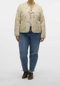 Beige gewatteerde jas met striksluiting, geborduurde details en twee voorkruiden. Gepaard met blauwe jeans en bruin veterschoenen.