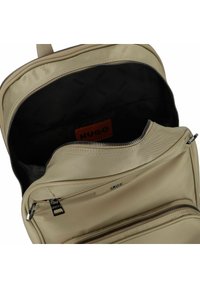 Mochila beige con un exterior suave y texturizado, que cuenta con un cierre de cremallera, un forro negro y una etiqueta de logo prominente en el interior.