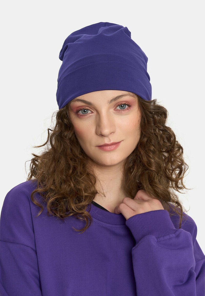 Fioletowy beret z miękkiego, teksturowanego materiału i luźnym krojem. Osoba nosi dopasowaną, fioletową, oversize'ową bluzę.