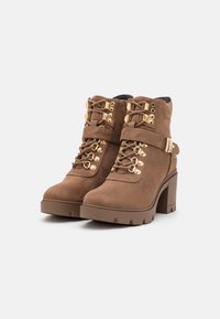 Stivali alla caviglia in suede marrone con hardware dorato, lacci frontali, dettaglio a fascia e tacco a blocco. Presentano una superficie testurizzata e una suola robusta.