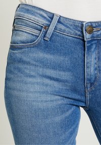 Vaqueros de mezclilla azul con un acabado desgastado. Cierre de botón con la marca "Lee". Diseño estándar de cinco bolsillos, costuras sutiles y trabillas para el cinturón.
