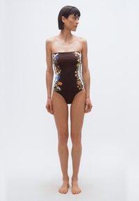 Γυναίκα με κοντά σκουρόχρωμα μαλλιά, που φοράει ένα bandeau καφέ μαγιό με πολύχρωμα λουλουδάτα και ζωικά μοτίβα, όρθια ξυπόλυτη μπροστά σε απλό φόντο.