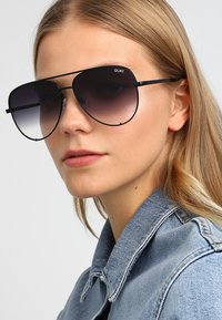 Lunettes de soleil aviateur noires avec des verres dégradés de foncé à clair ; design minimal avec des branches fines ; modèle une veste en denim bleu.