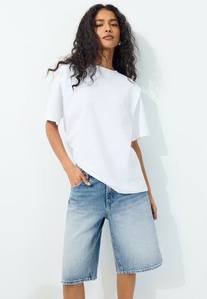 Frau mit langen lockigen Haaren, die ein schlichtes weißes T-Shirt und hellblaue, knielange Jeansshorts trägt und mit einer Hand in der Tasche steht.