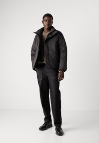 Giacca a vento nera con colletto alto, tessuto texturizzato e chiusura con zip. Indossata sopra un maglione di lana beige e abbinata a pantaloni di jeans scuri e scarpe nere.