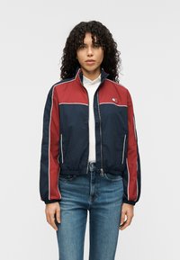 Μπλε ναυτικό και κόκκινο cropped μπουφάν από ελαφρύ υλικό, με λευκές ραφές και φερμουάρ στο κλείσιμο. Λογότυπο στο αριστερό στήθος.