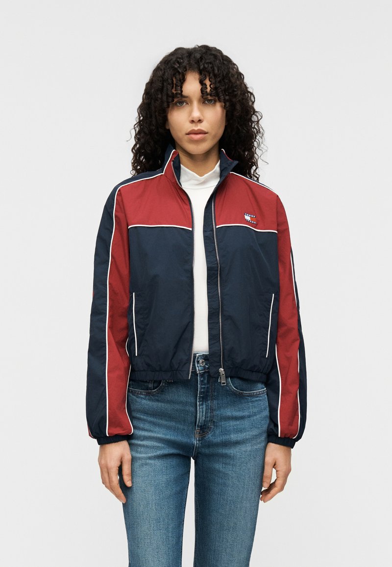 Μπλε ναυτικό και κόκκινο cropped μπουφάν από ελαφρύ υλικό, με λευκές ραφές και φερμουάρ στο κλείσιμο. Λογότυπο στο αριστερό στήθος.