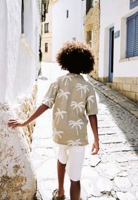 Camicia a maniche corte con motivo di palme in beige chiaro, abbinata a pantaloncini bianchi. Persona che cammina su una strada lastricata di ciottoli.