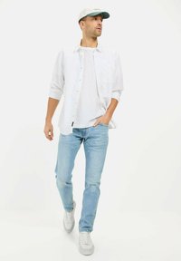 Biała koszula na guziki noszona na białym t-shircie, jasnoniebieskie dżinsy i białe trampki. Czapka jest jasnoszara z ciemniejszym daszkiem.