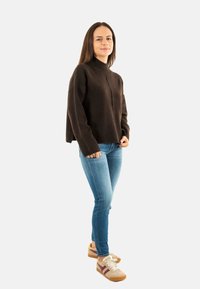 Maglione marrone a maglia con collo alto, orlo a coste, abbinato a jeans blu chiaro e sneakers beige con accenti bordeaux.