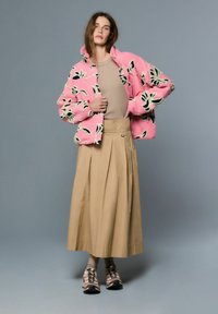 Roza, plišasta jakna s črnimi in kremnimi cvetličnimi vzorci, kombinirana z bež, naborkasto krilo in večbarvnimi športnimi čevlji.