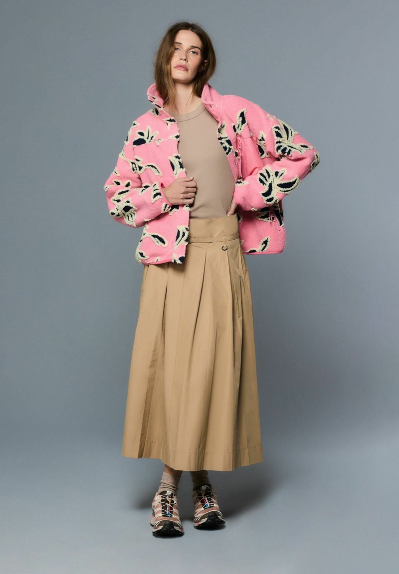 Roza, plišasta jakna s črnimi in kremnimi cvetličnimi vzorci, kombinirana z bež, naborkasto krilo in večbarvnimi športnimi čevlji.