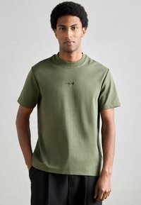Īss piedurkņu t-krekls olīvu zaļā krāsā no mīksta auduma, ar mazu melnu logotipu krūšu centrā un apaļu kakla izgriezumu.