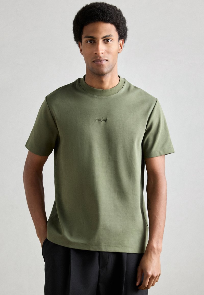 Īss piedurkņu t-krekls olīvu zaļā krāsā no mīksta auduma, ar mazu melnu logotipu krūšu centrā un apaļu kakla izgriezumu.
