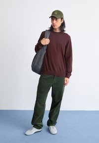 Brauner Pullover, grüne Cordhosen, graue Tasche und grüne Mütze mit Logo. Das Model steht auf einem blauen Boden vor einem hellen Hintergrund.