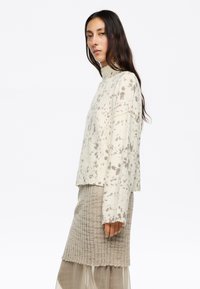 Maglione lavorato a maglia di colore chiaro con un collo alto, caratterizzato da un motivo a macchie. Abbinato a una gonna beige texture a coste.