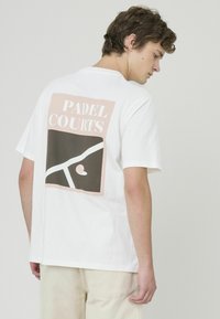 Jeune homme de profil portant un T-shirt blanc avec un grand motif et le texte "Terrains de Padel" dans le dos.