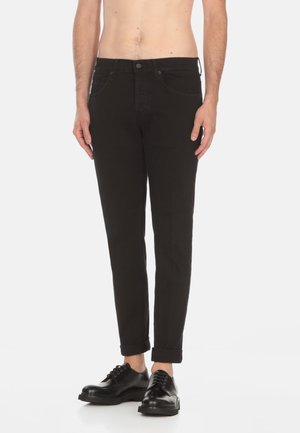 Mężczyzna ubrany w czarne dopasowane jeansy z podwiniętymi nogawkami i czarne skórzane buty, stoi na białym tle.