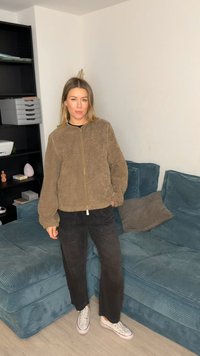 Καφέ, αφράτο τζάκετ με φερμουάρ και φουσκωτά μανίκια· μαύρο παντελόνι· λευκά αθλητικά παπούτσια. Καναπές και ράφια με διάφορα αντικείμενα στο φόντο.