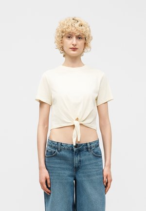 Ung person med kort, krøllet blondt hår iført en cremefarvet kortærmet cropped top bundet ved taljen og blå højttaljede jeans.