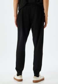 Schwarze Sweatpants mit elastischem Bund, schmal zulaufenden Beinen und Bündchen. Glatte Textur, keine sichtbaren Muster. Getragen mit weißen Sneakers.