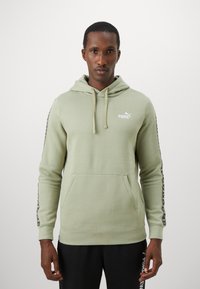 Mand iført en lysegrøn Puma hættetrøje med logo på brystet og mønstrede striber på ærmerne, sammen med sorte Puma joggingbukser.