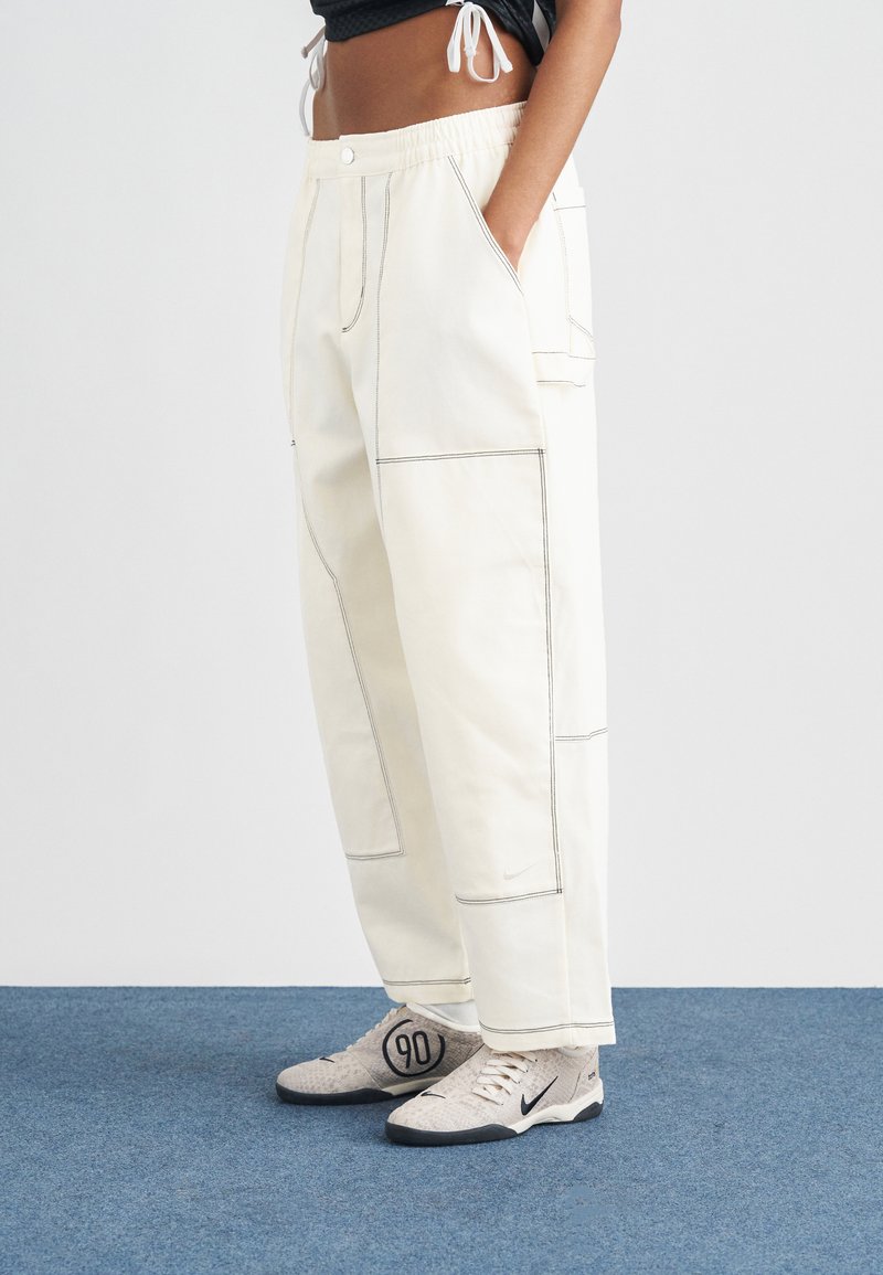 Λευκό υφασμάτινο παντελόνι με ορατές ραφές, πλαϊνές τσέπες και ελαστική μέση· συνδυάζεται με γκρι παπούτσια Nike με κυκλικό λογότυπο.