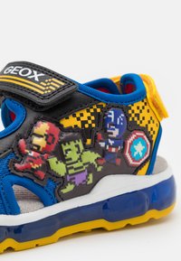 Melna un dzeltena sandele ar zilo akcentu, kurā attēloti pikselīto supervaroņu tēli. Ar Velcro siksniņu un mīkstu zoli komfortam.