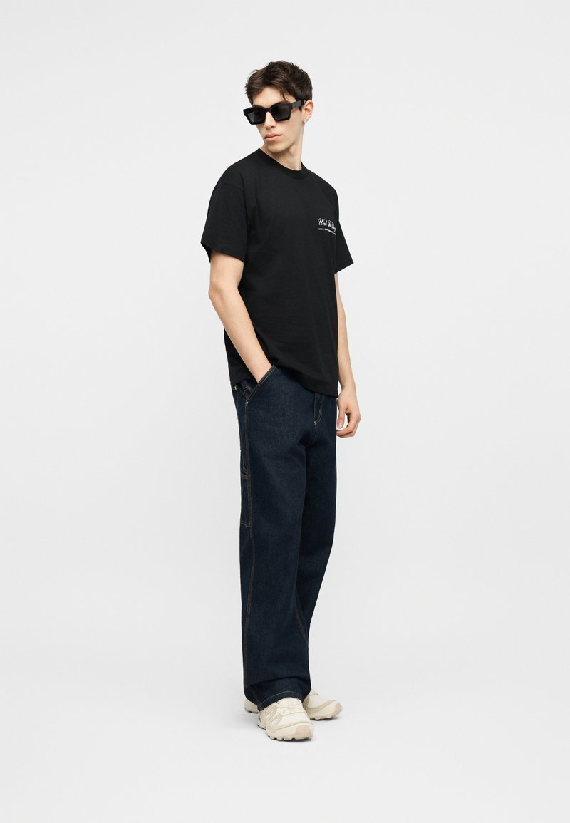 Giovane uomo con occhiali da sole neri, t-shirt nera con grafica, jeans larghi e scuri e sneakers bianche, in piedi con una mano in tasca.