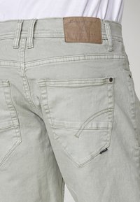 Svetlo sivi traper hlače, pogled od zadaj, ki prikazuje dva zadnja žepa, zanke za pas in rjav usnjen logotip na pasu.