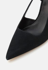 Black suede high-heeled shoe with a pointed toe and cutout design, featuring a smooth interior and a reflective finish on the sole.