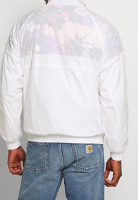 Giacca bianca leggera con retro semi-trasparente caratterizzato da un motivo floreale. Presenta polsini elastici e vita elasticizzata, abbinata a jeans blu.