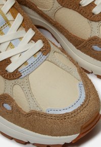 Teniska s hnědými a béžovými semišovými a koženými panely, bílými tkaničkami, žlutými a světle modrými akcenty a bílou gumovou podrážkou.