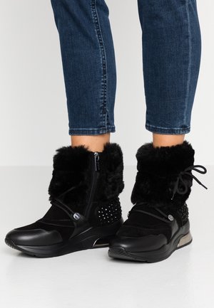 Osoba nosząca czarne botki z futrzaną wyściółką, zamkami błyskawicznymi, ćwiekami i sznurowadłami, zestawione z krótkimi, ciemnoniebieskimi dżinsami na białej podłodze.