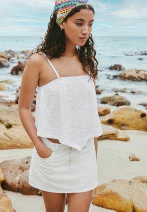 Junge Frau mit lockigem Haar, die ein bunt gehäkeltes Stirnband, ein weißes ärmelloses Oberteil und einen weißen Rock trägt und an einem felsigen Strand steht.