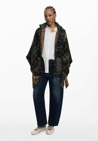 Übergröße, dunkelgrünes Karo-Poncho mit braunen Akzenten, getragen über einem weißen Hemd, kombiniert mit dunklen Jeans und hellen Schuhen.