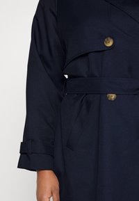 Trench coat blu navy scuro con chiusura a bottone, ampi rever e una cintura abbinata. Il tessuto ha una superficie liscia e le maniche hanno polsini arrotolati.