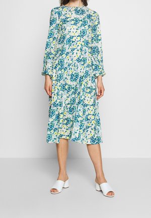 Femme portant une robe fleurie à manches longues, longueur genou, avec des fleurs bleues et jaunes, assortie à des chaussures blanches à talons ouverts.