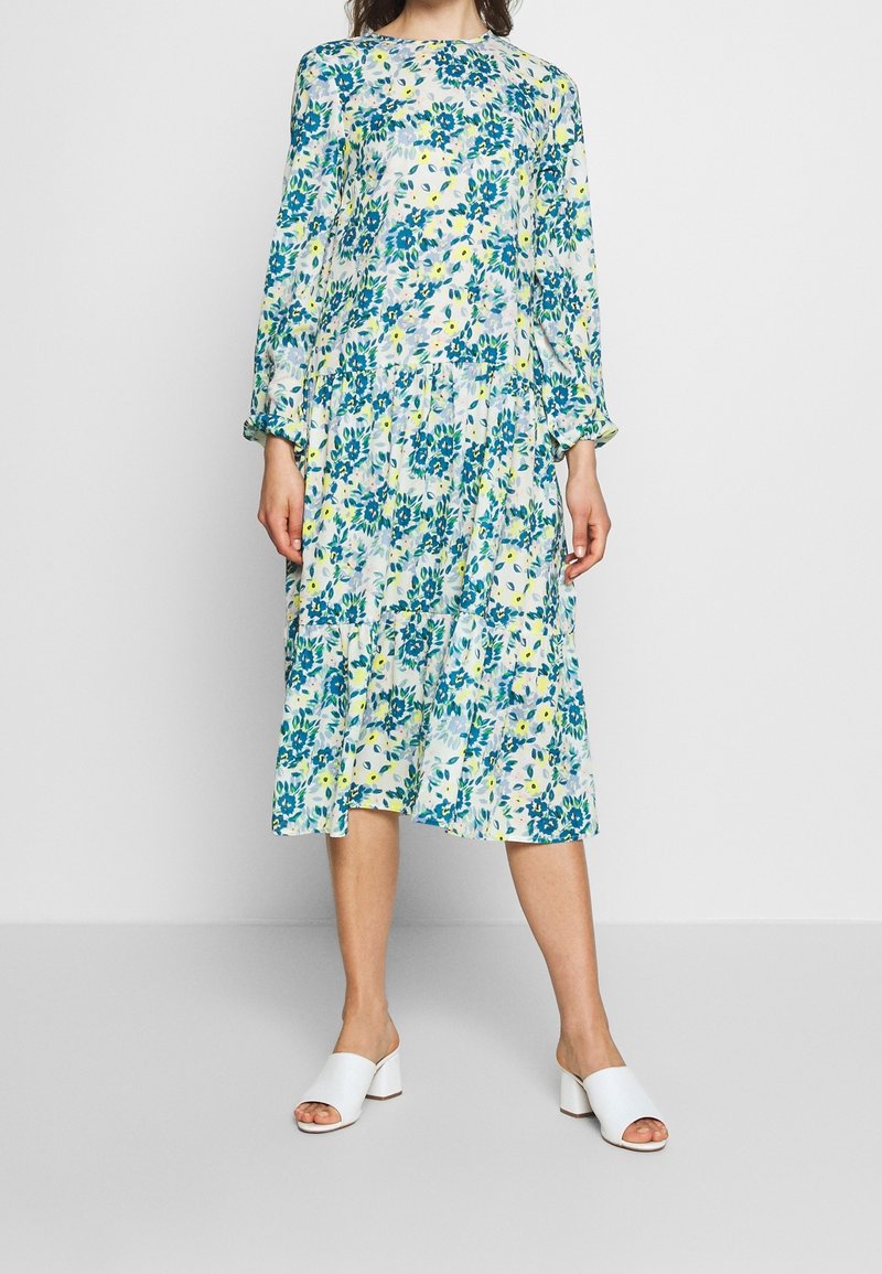 Femme portant une robe fleurie à manches longues, longueur genou, avec des fleurs bleues et jaunes, assortie à des chaussures blanches à talons ouverts.