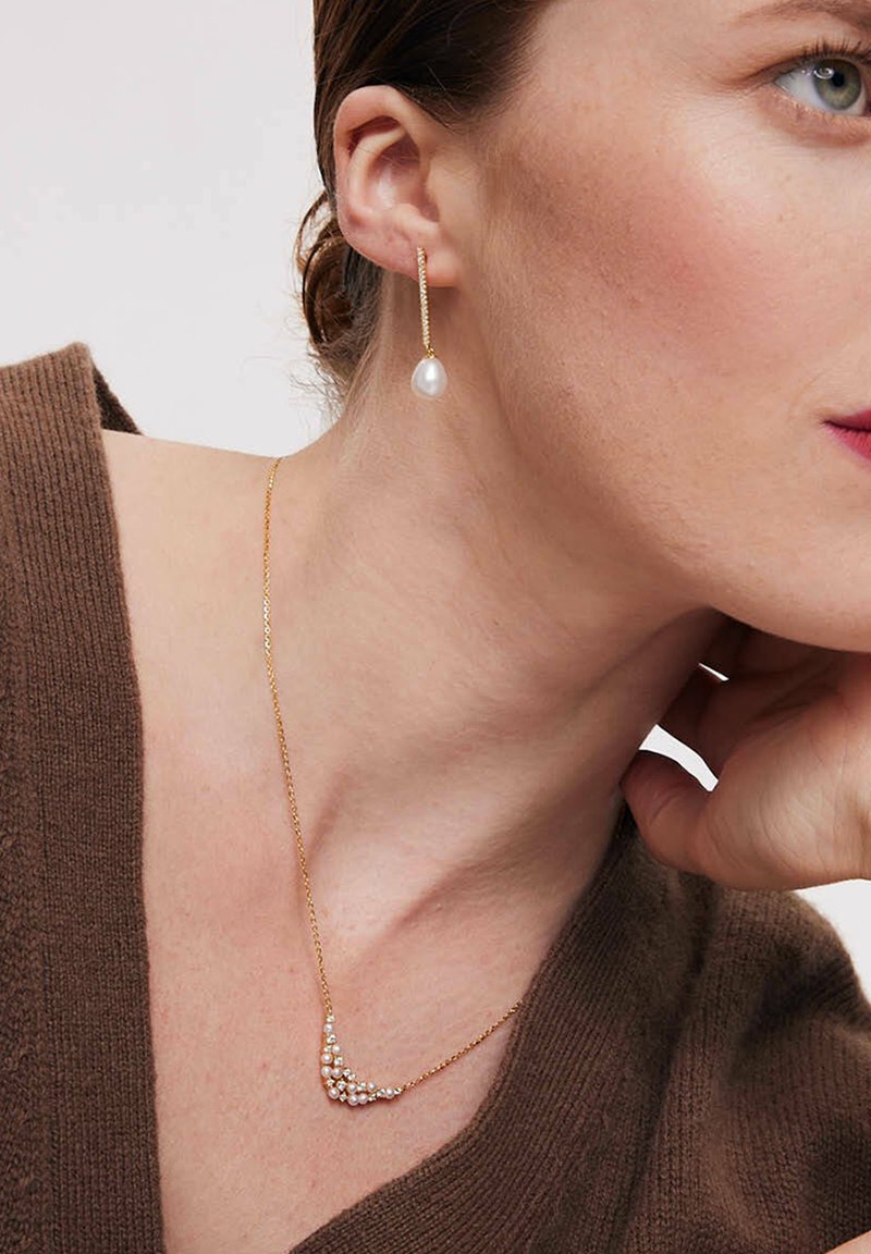 Złoty naszyjnik o zakrzywionym kształcie, zdobiony małymi, błyszczącymi koralikami. Proste złote kolczyki w kształcie kropli z akcentem w postaci białej perły.