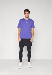 Mand, der står op ad en enkel væg iført en lilla kortærmet skjorte, sorte shorts, sorte leggings og hvide træningssko.