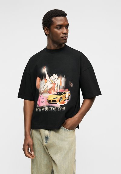 Άνδρας που φοράει μαύρο υπερμεγέθες μπλουζάκι με γραφικά μιας καρτουνίστικης γυναίκας, ενός κίτρινου σπορ αυτοκινήτου, μιας τσάντας αγορών και το κείμενο "WWW.GCDS.COM".