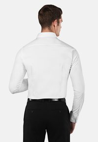 Chemise blanche à manches longues avec un col classique, texture lisse et boutons de poignet, associée à un pantalon noir doté de deux poches arrière.