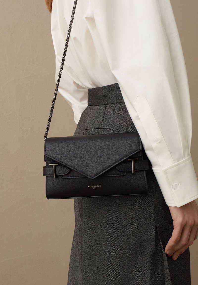 Sac à main en cuir noir avec un design structuré, doté d'un rabat d'enveloppe, de quincaillerie en argent et d'une chaîne en bandoulière. Surface texturée et marquage subtil.