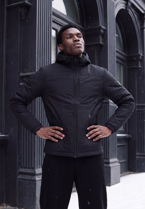 Homme en veste noire debout, les mains sur les hanches, devant un bâtiment sombre alors que la neige tombe.