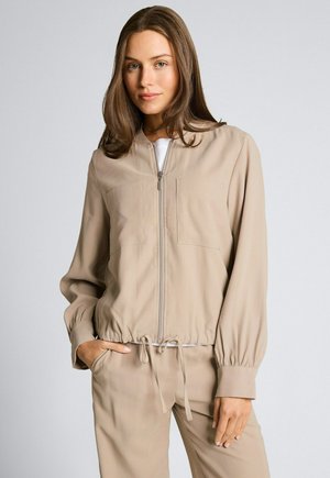 Kobieta o prostych, brązowych włosach, ubrana w beżową kurtkę na zamek i dopasowane spodnie, stojąca z jedną ręką w kieszeni na jasnym tle.