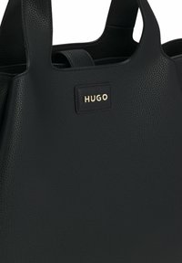 Sort lædertaske med tekstureret finish, forsynet med en guldfarvet "HUGO" logo-plade. To robuste håndtag til bæring.